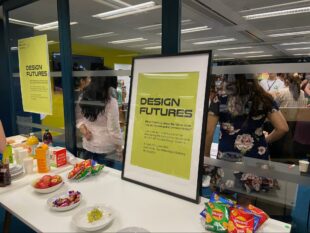 A yellow poster in a black frame. The poster is titled “Design Futures”. It is placed on a table where there are snacks and refreshments, with people standing around behind it.