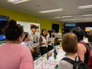 Students presenting their projects around a table, on the table are some maps with markers. GDS staff are chatting with the students.