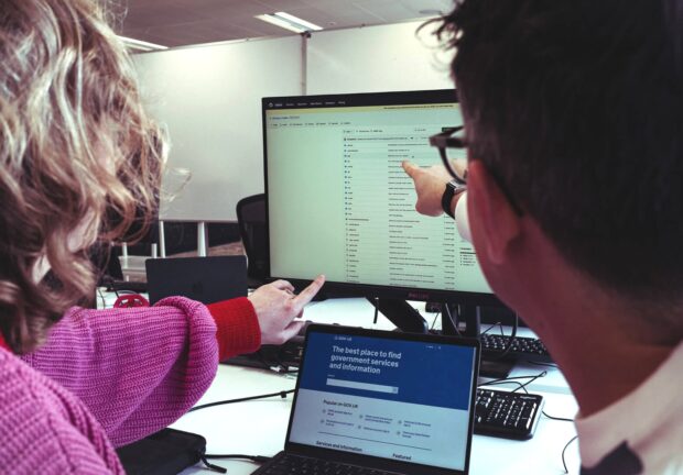 Two people sitting in front of a laptop while pointing at a larger computer monitor. The laptop shows the GOV.UK homepage and the monitor shows the archived Static GitHub repository page.
